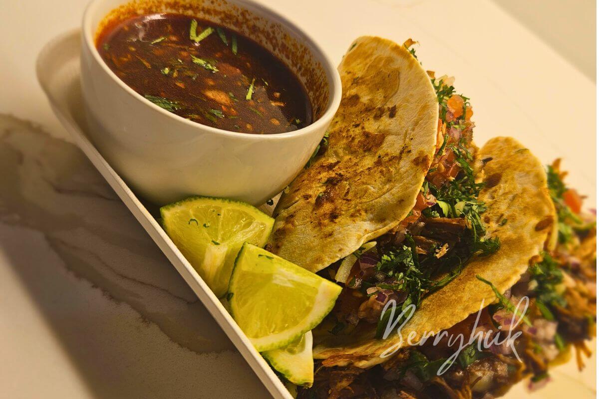 Birria tacos served with consommé, lime wedges, and fresh cilantro on a plate.
