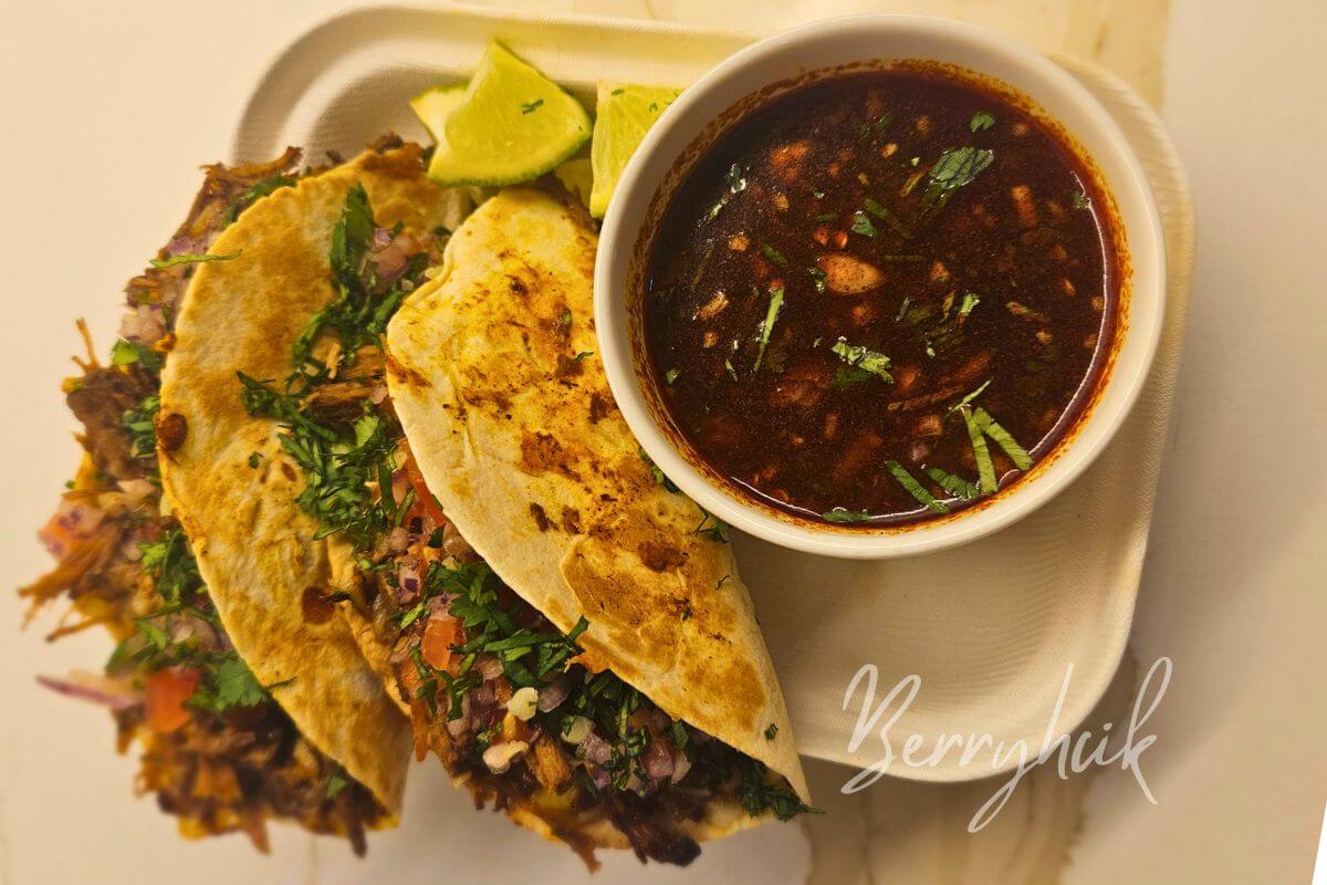 Birria taco is ready to be dipped into consommé, showing the plled beef and glossy red broth.