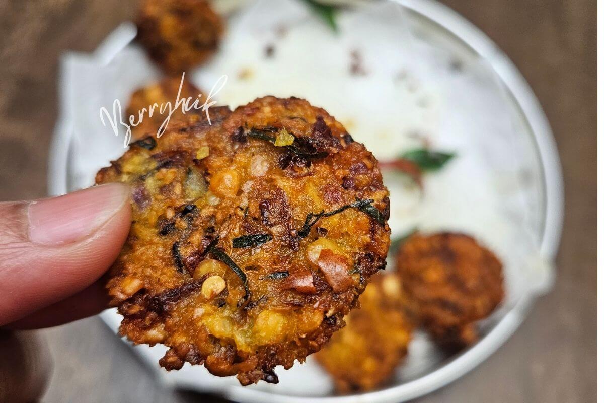 Hand holding a crispy Sri Lankan parippu vada above a bowl of rice, showing its golden, crunchy texture.