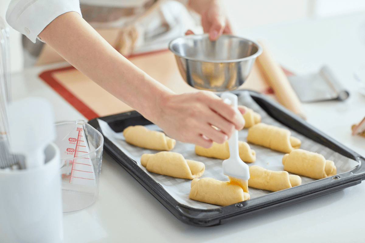 Person pouring batter onto a tray of crescent‑shaped pastries in a bright kitchen